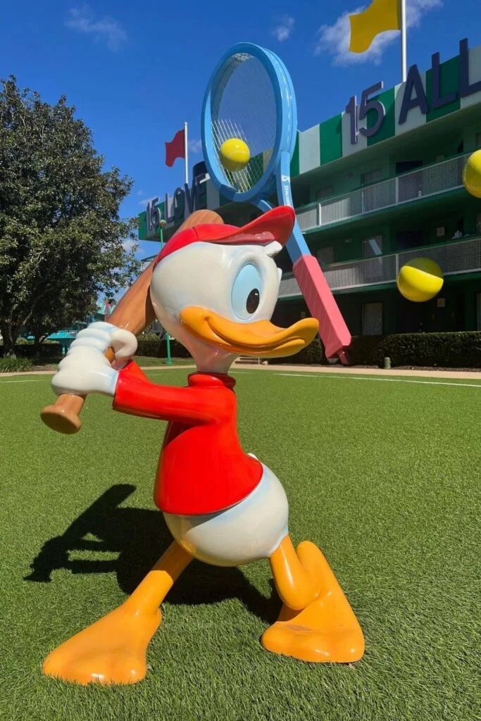 Photo of a life-sized Huey statue holding a baseball bat with the Center Court buildings at Disney's All-Star Sports Resort in the background.
