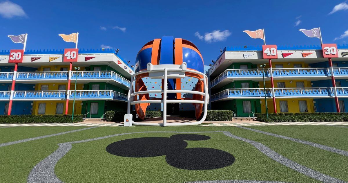 Horizontal photo of a 3-story blue and orange football helmet nestled between two hotel buildings. In the foreground is a faux football field with a large Mickey Mouse head painted in the middle.