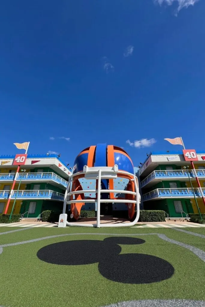 Vertical photo of a 3-story blue and orange football helmet nestled between two hotel buildings. In the foreground is a faux football field with a large Mickey Mouse head painted in the middle.