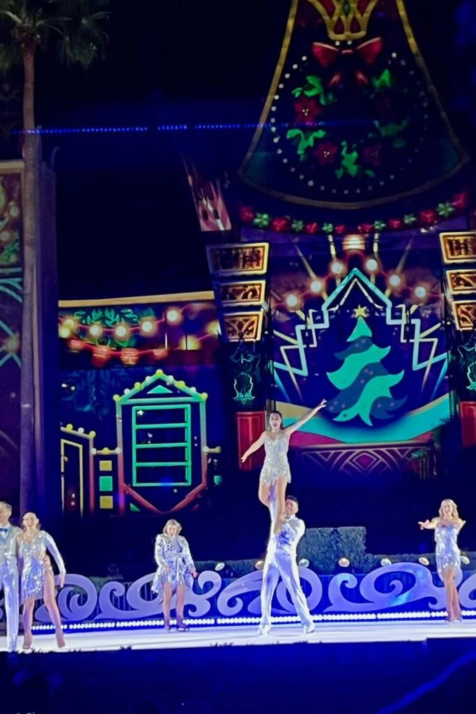 Photo of a man on ice skates lifting a woman during a scene from the Glisten! ice skating show at Jollywood Nights. Beside them are other men and women on ice skates wearing silver sparkly costumes.