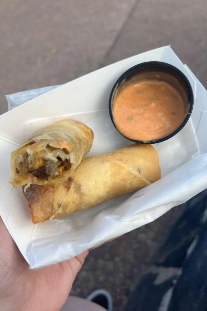 Photo of a basket with 2 cheeseburger spring rolls with a cup of burger dipping sauce.