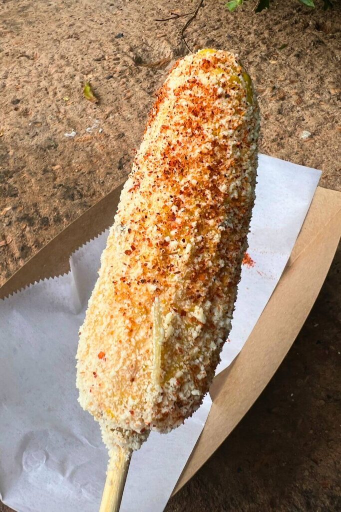 Closeup of elote, corn on the cob covered in chipotle aioli, queso cotija, and Taijin chili-lime powder, on a stick.