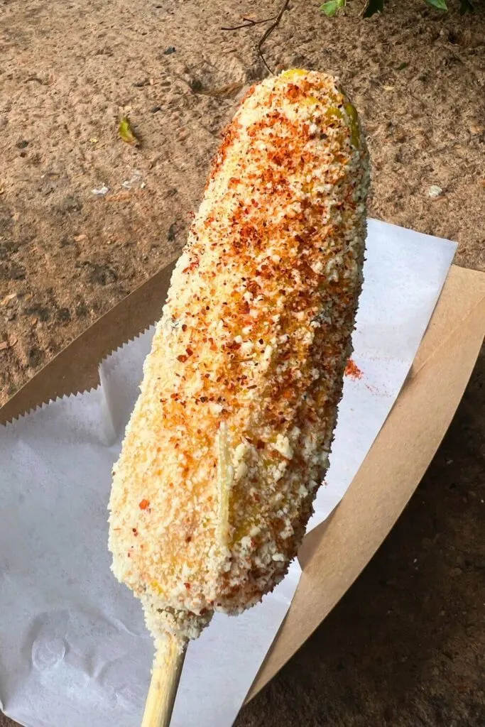 Closeup of elote, corn on the cob covered in chipotle aioli, queso cotija, and Taijin chili-lime powder, on a stick.