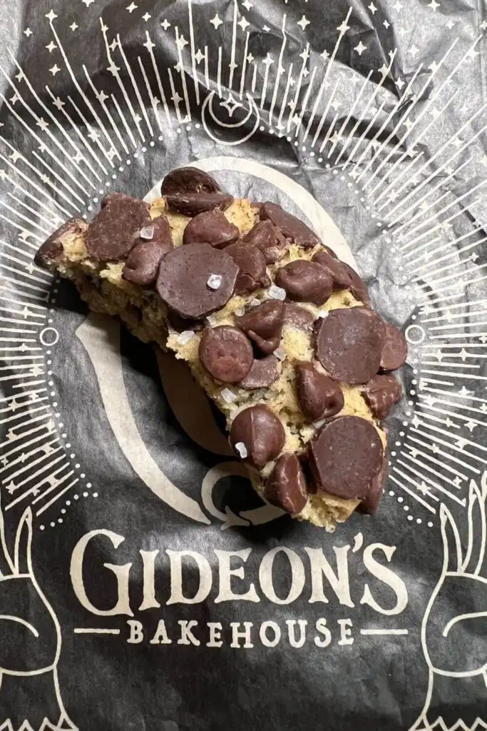 Photo of half of a chocolate chip cookie from Gideon's Bakehouse on top of a paper with the bakery's logo on it.