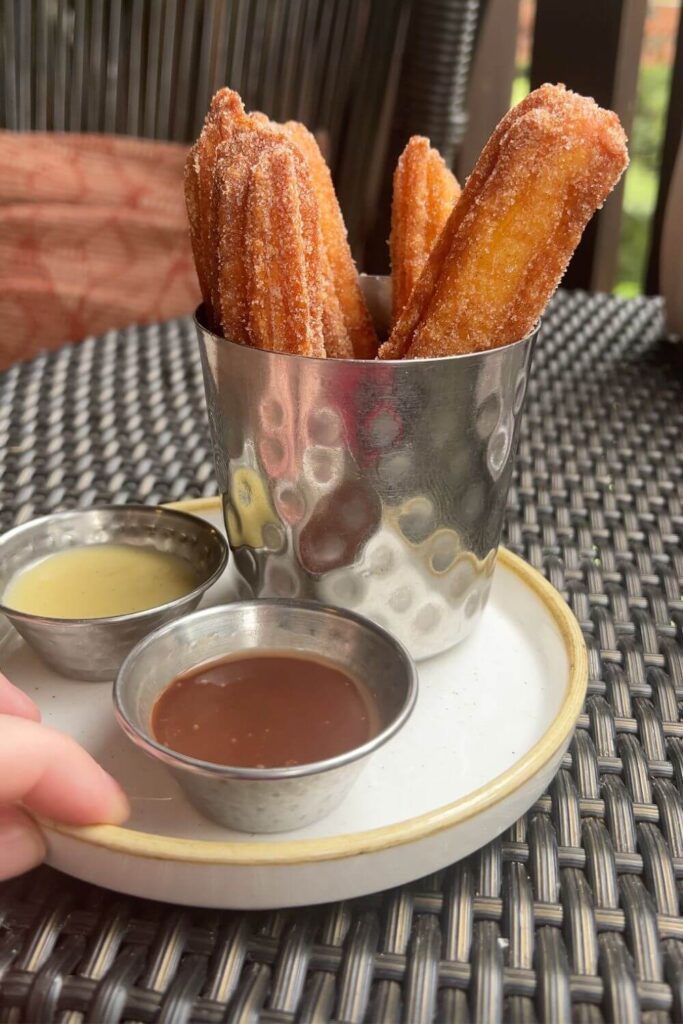 Photo of freshly fried churros in a metal cup with 2 small dipping cups filled with vanilla and chocolate sauce.