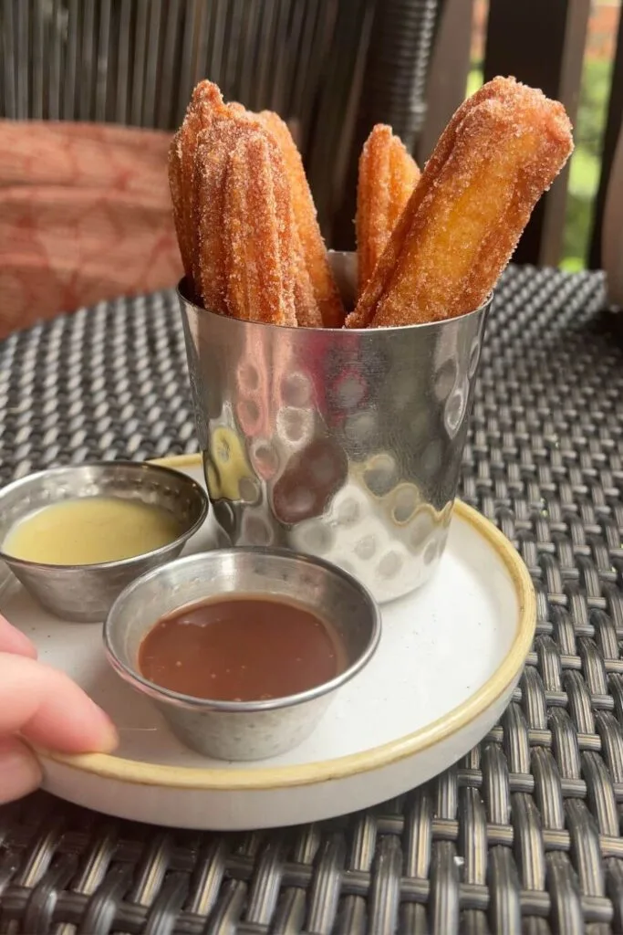 Photo of freshly fried churros in a metal cup with 2 small dipping cups filled with vanilla and chocolate sauce.