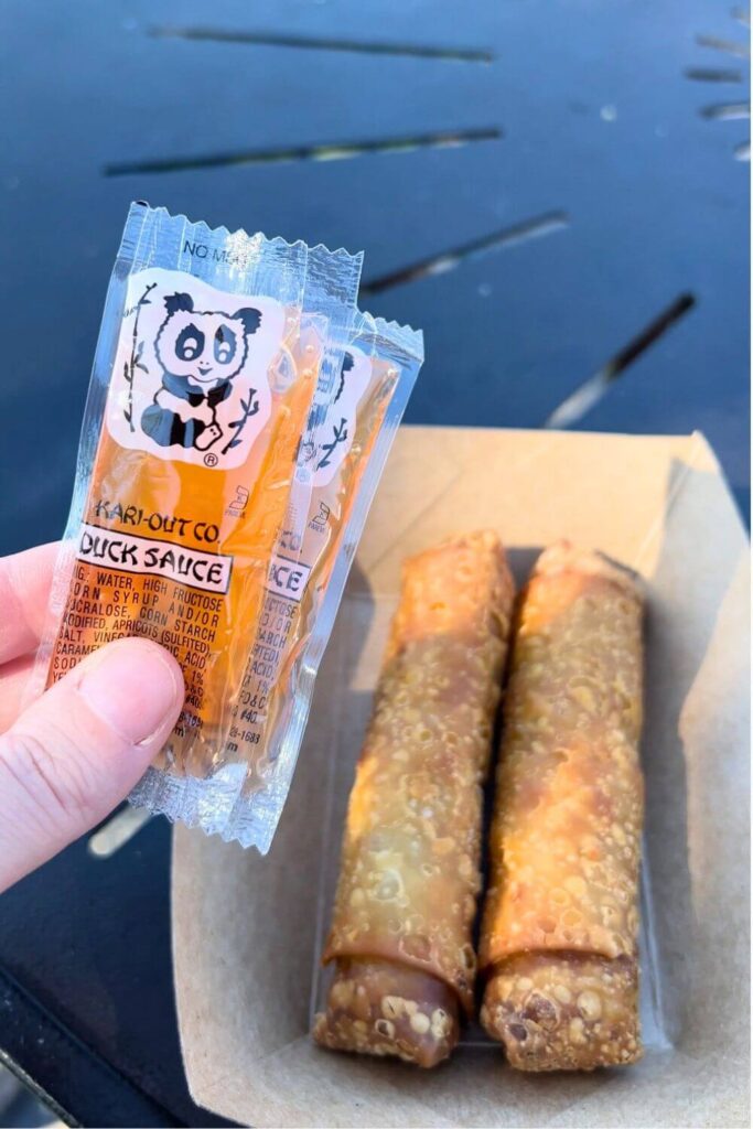 Photo of 2 crispy fried pork egg rolls in a basket, with a hand holding 2 packets of duck sauce.