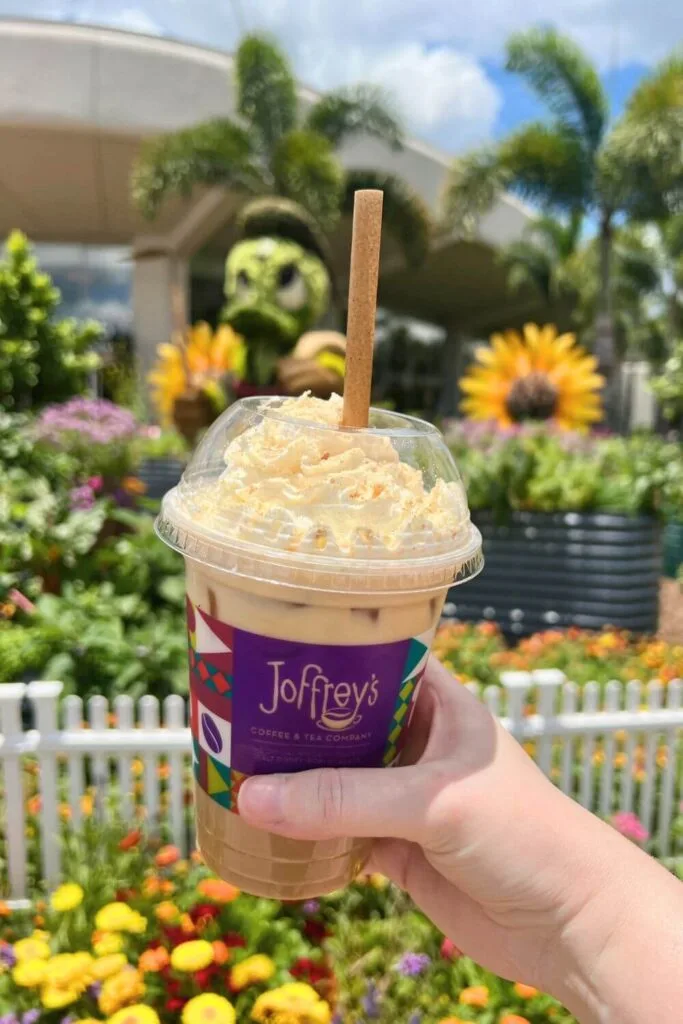 Closeup of an iced seasonal latte from Joffrey's in World Discovery at Epcot, with a Donald Duck topiary in the background.