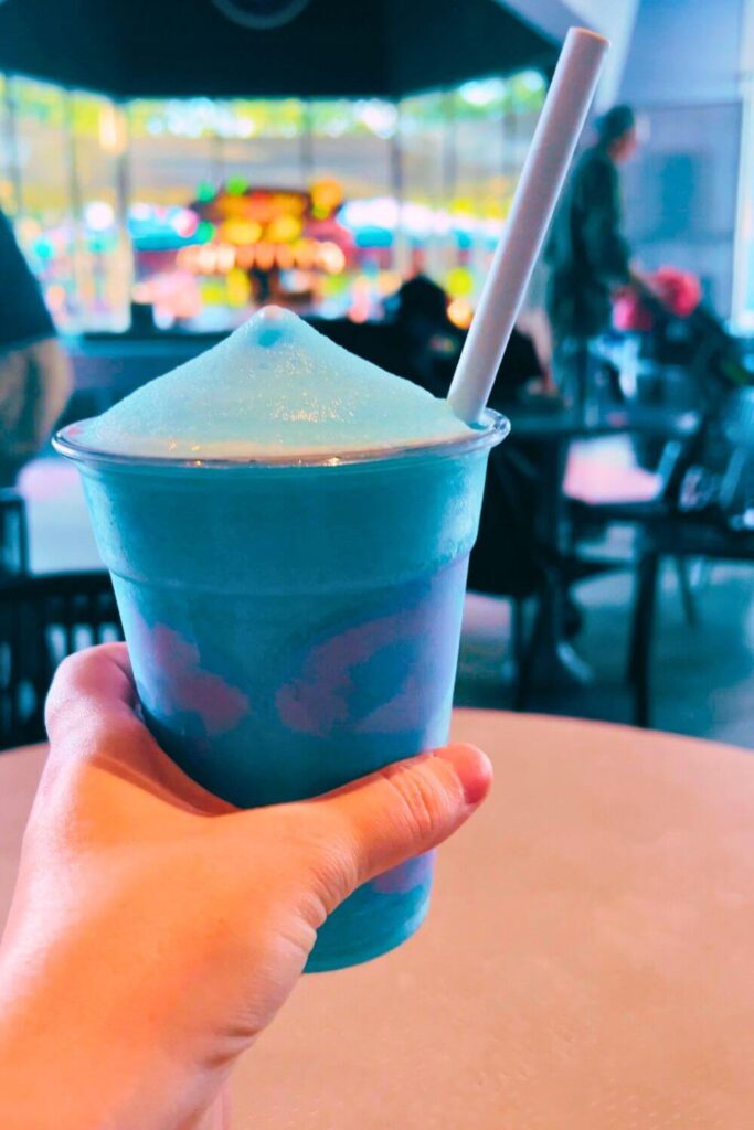 Closeup of a hand holding a cup of Sulley Slush, a blue raspberry slush with purple whipped cream dots on the wall of the cup.