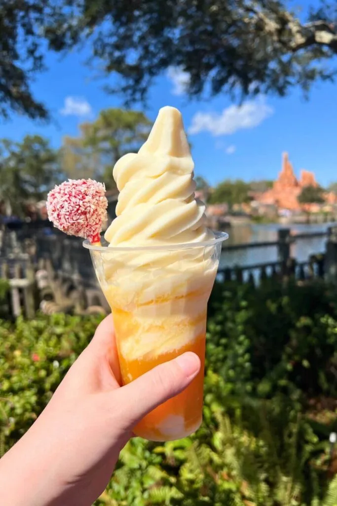 Photo of the tropical serenade Dole whip float from Magic Kingdom, featuring coconut soft serve in a cup of POG juice, topped with a pineapple cake pop.